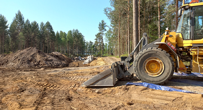 Påbörjat bygge av gator och villatomter i Södra Hovmantorp. På bilden syns en gul grävmaskin, sandmark och skog.