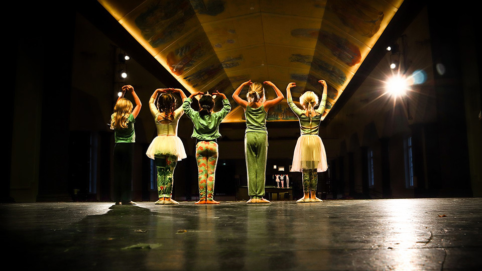 Foto bakifrån på fem barn som står på en scen och har olika danspositioner.