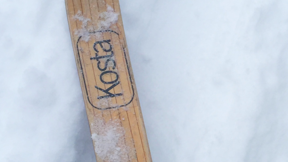 Syntolkning: En bild på snö med en närbild på en del av en skida som ligger i mitten på bilden. På den träfärgade gulaktiga skidan finns texten "Kosta" i svart tunn färg.